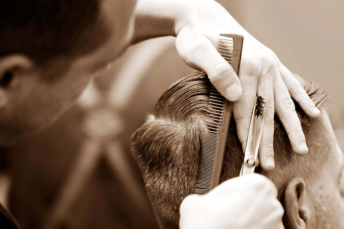 A close-up of a barber giving a haircut to a man, using a comb and scissors to carefully trim the hair on the side of his head.
