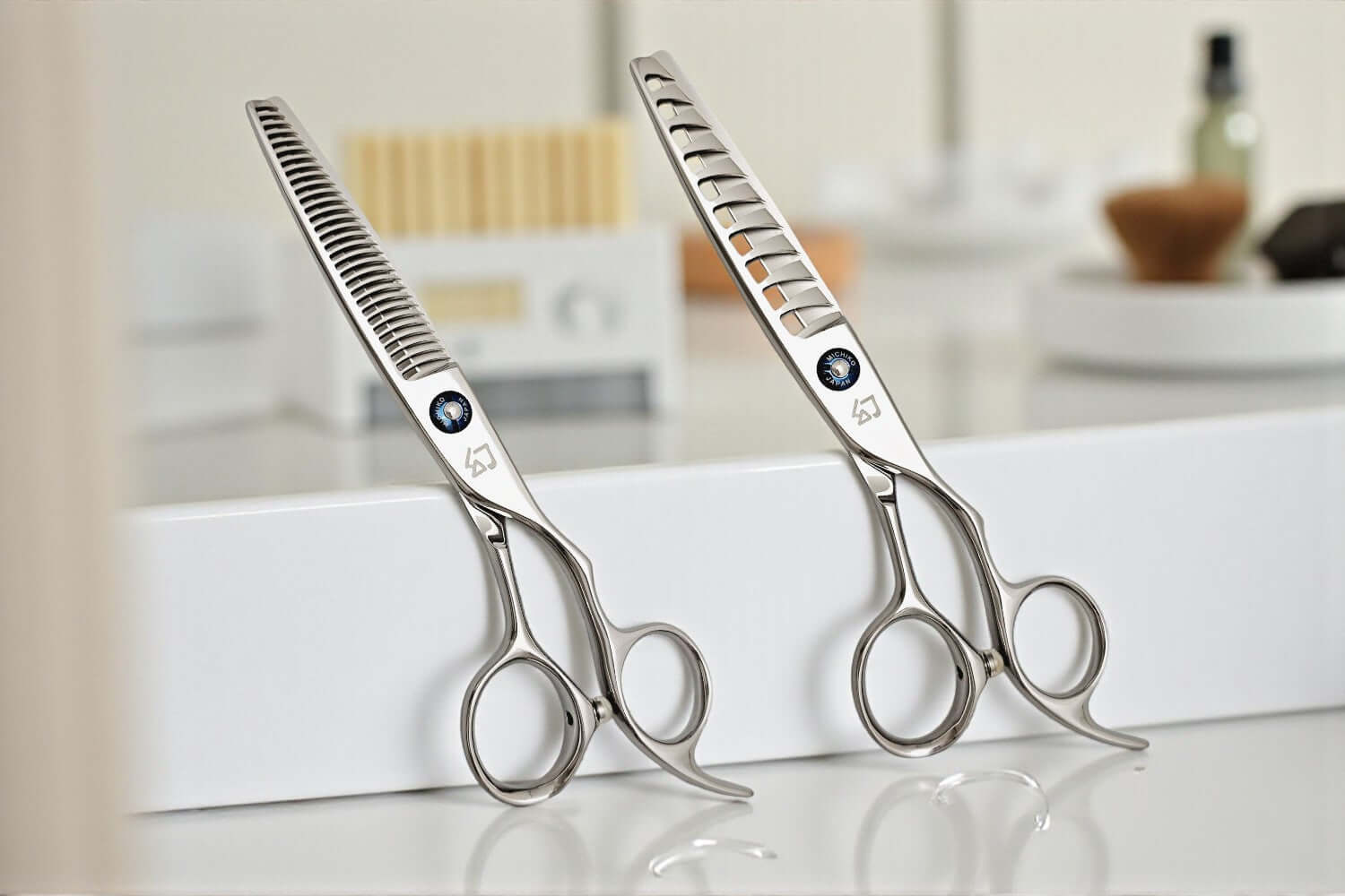 Two pairs of silver professional hair scissors (one texturizing and one thinning shear) with blue jewel accents, balanced on a white salon station.