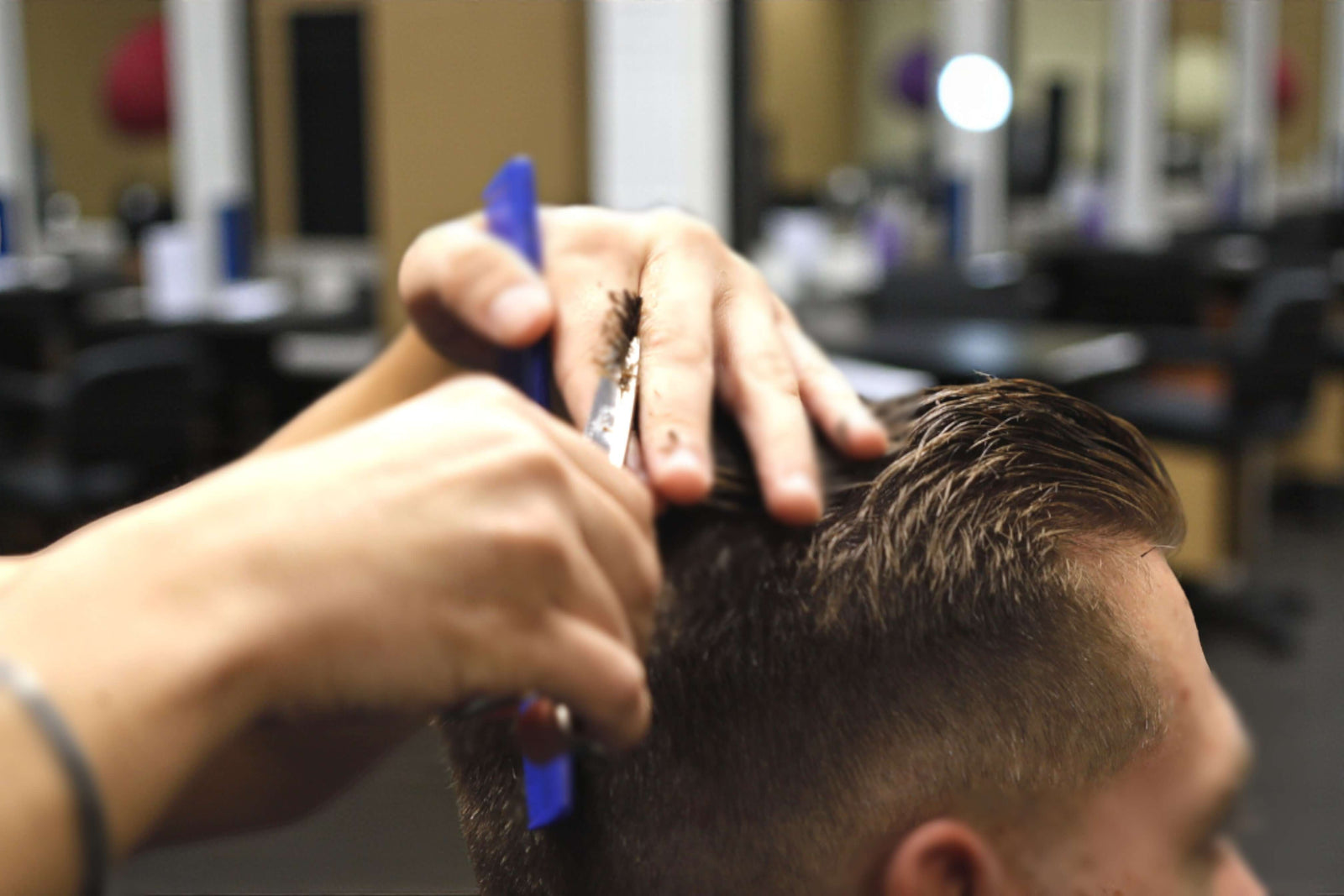 A close-up of a barber's hands holding a blue comb and scissors, actively trimming the slicked-back top of a man's fade haircut.