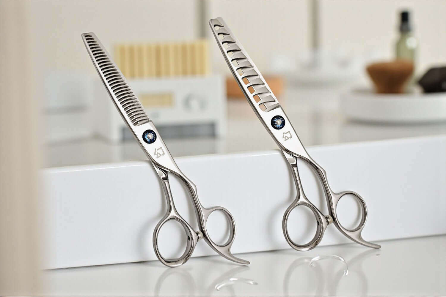 Two pairs of silver professional hair scissors (one texturizing and one thinning shear) with blue jewel accents, balanced on a white salon station.