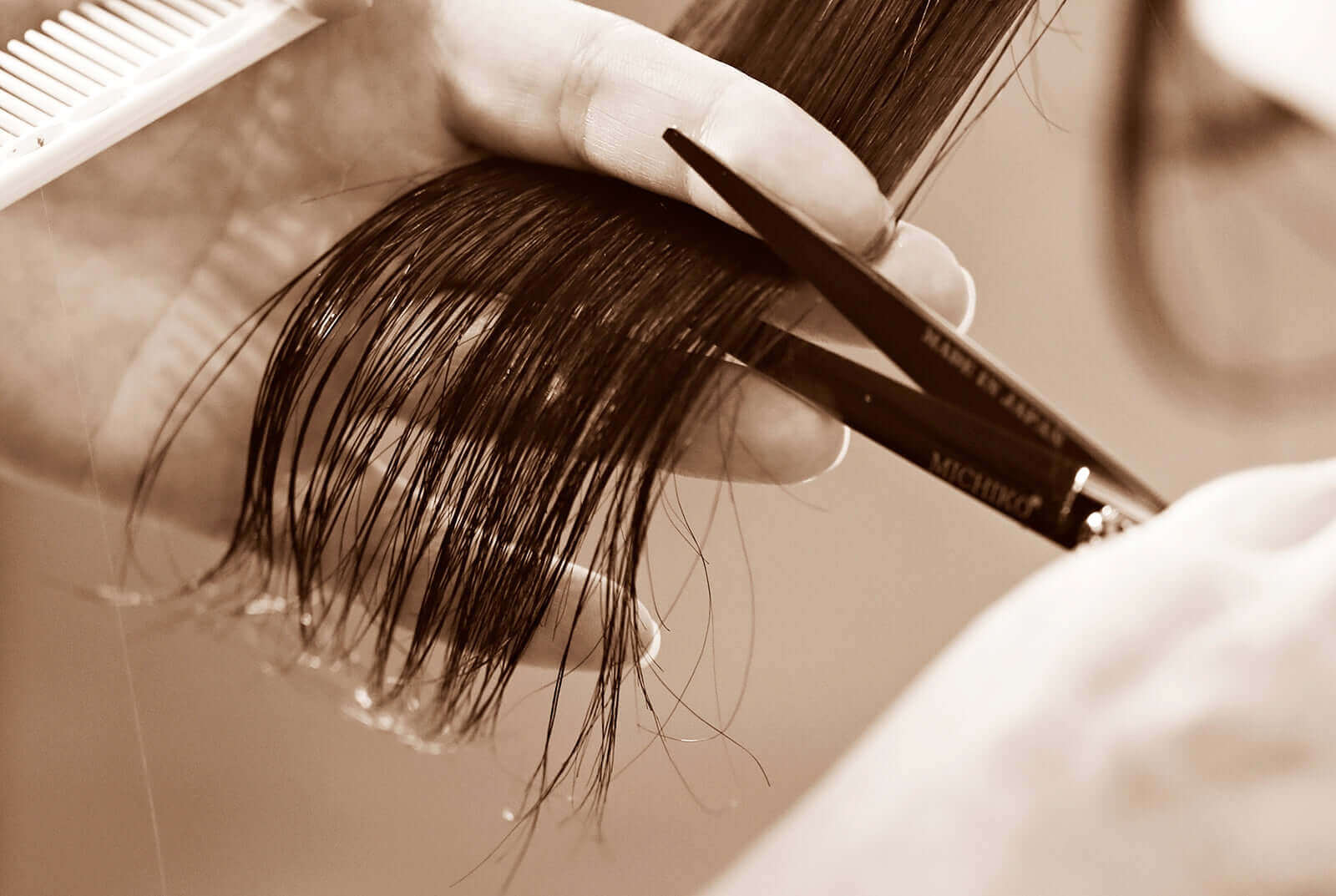 A sepia-toned close-up of a hairstylist cutting wet hair with professional scissors, holding a section between their fingers while a comb is visible in the background.