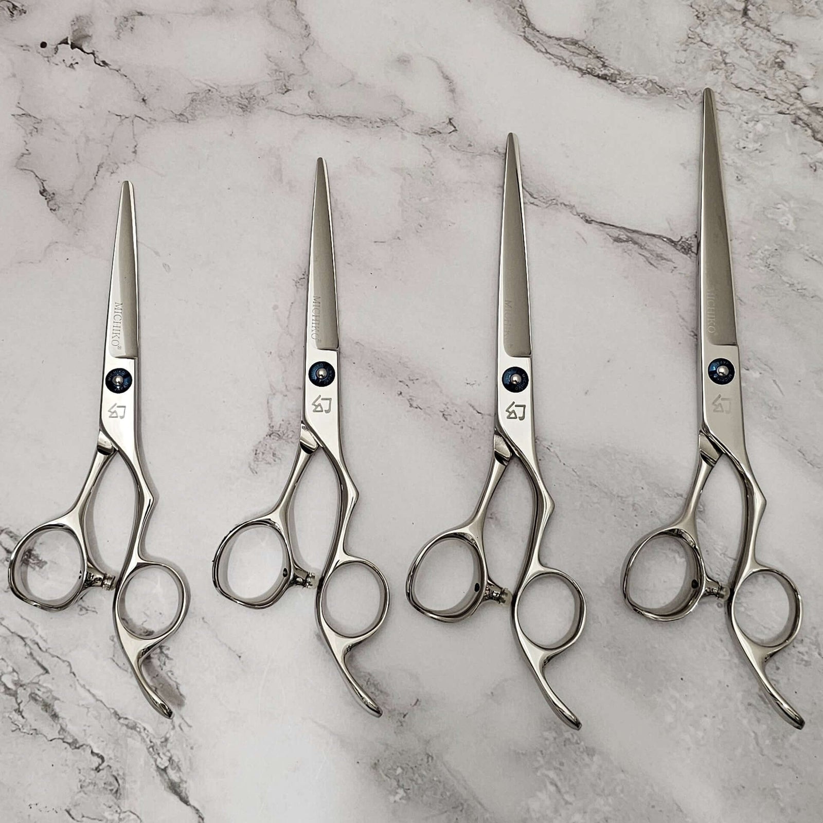 Four professional stainless-steel hair cutting shears arranged side by side on a light marble surface, showcasing ergonomic handles and precision blades