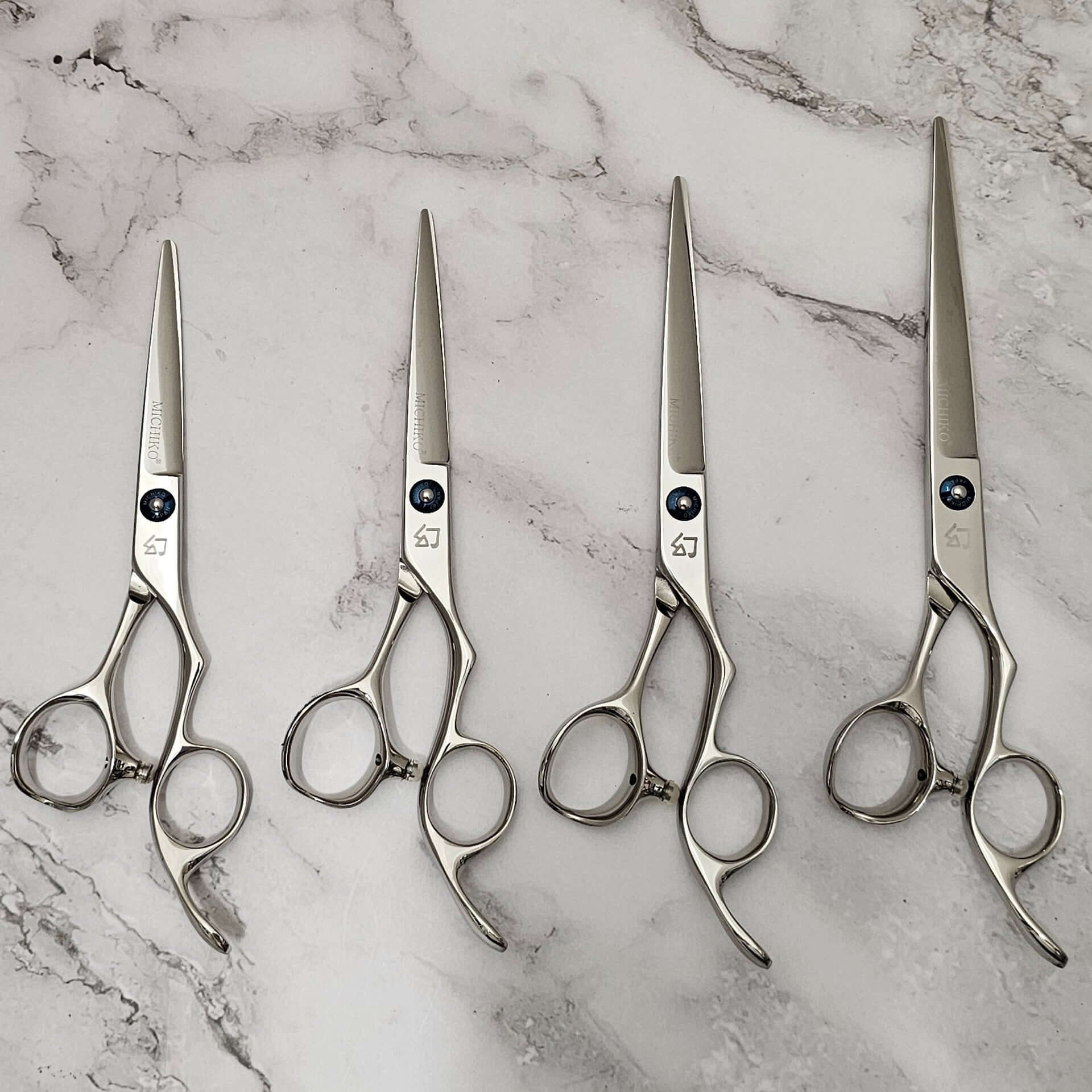 Four professional stainless-steel hair cutting shears arranged side by side on a light marble surface, showcasing ergonomic handles and precision blades