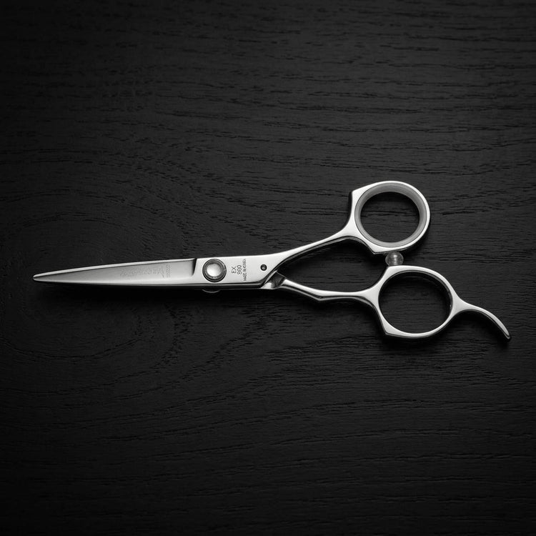 A pair of polished stainless steel hairdressing shears with ergonomic loop handles, displayed diagonally on a dark, textured wooden surface.