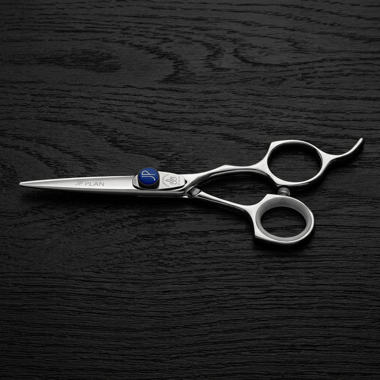 A pair of silver professional hair shears with a small blue accent screw, displayed diagonally on a dark wood-textured background.