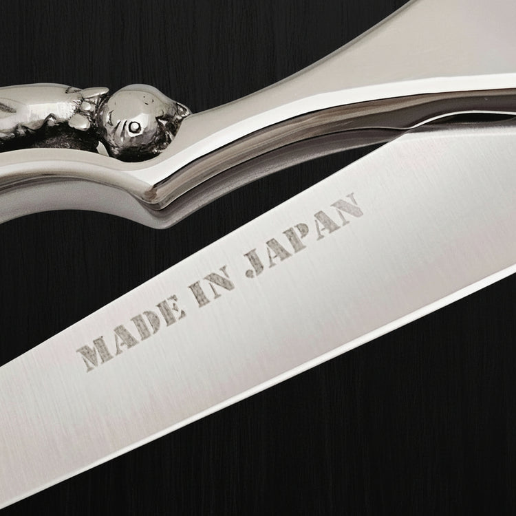 Close-up of an engraved kitten on a handle of hair shears with 'MADE IN JAPAN' engraving on the blade on a black oak textured background