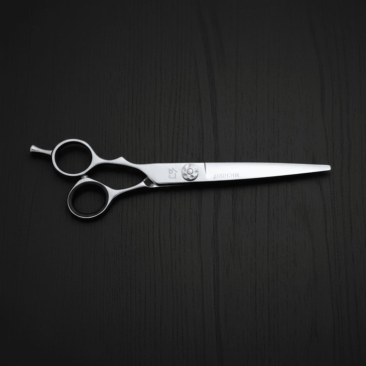 A pair of silver metal lefty hair shears displayed horizontally on a dark wood-textured background.
